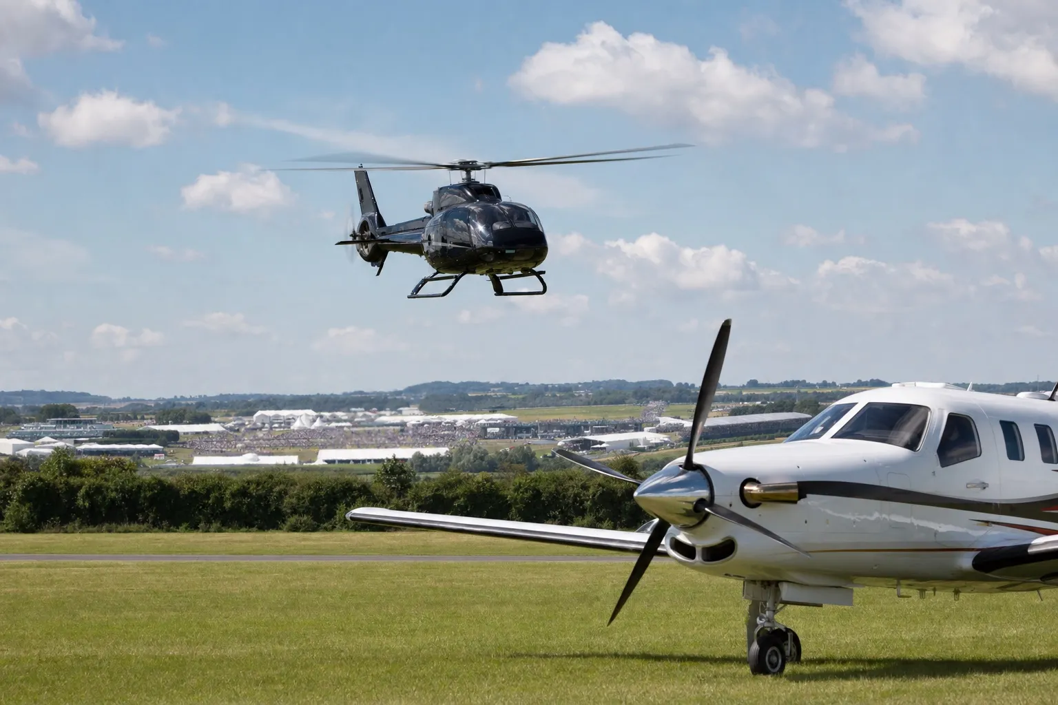 A helicopter and a turboprop aircraft at a grass airfield near Silverstone on British Grand Prix weekend with crowds and grandstands in the distance