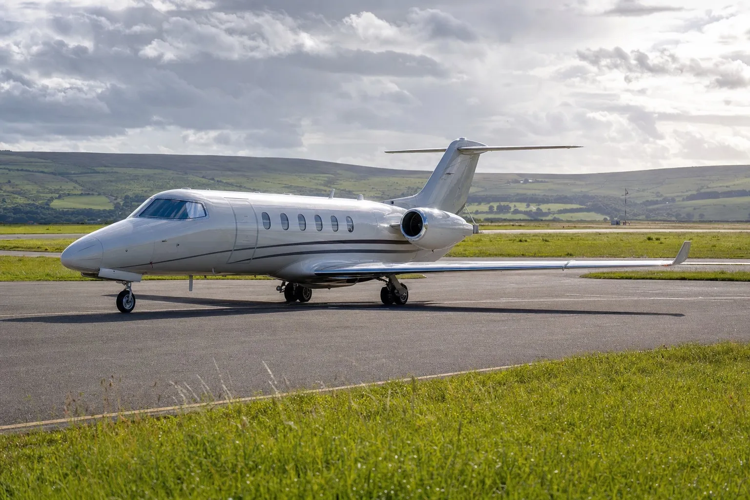 A light private jet on the apron of a small regional UK airport with green hills behind it, under a bright cloudy northern sky