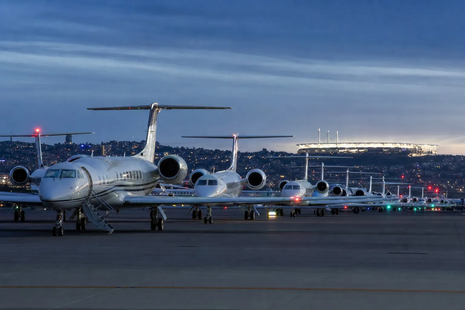 Private jets and charter aircraft parked in long rows at a European airport at dusk with a floodlit stadium glowing in the distance