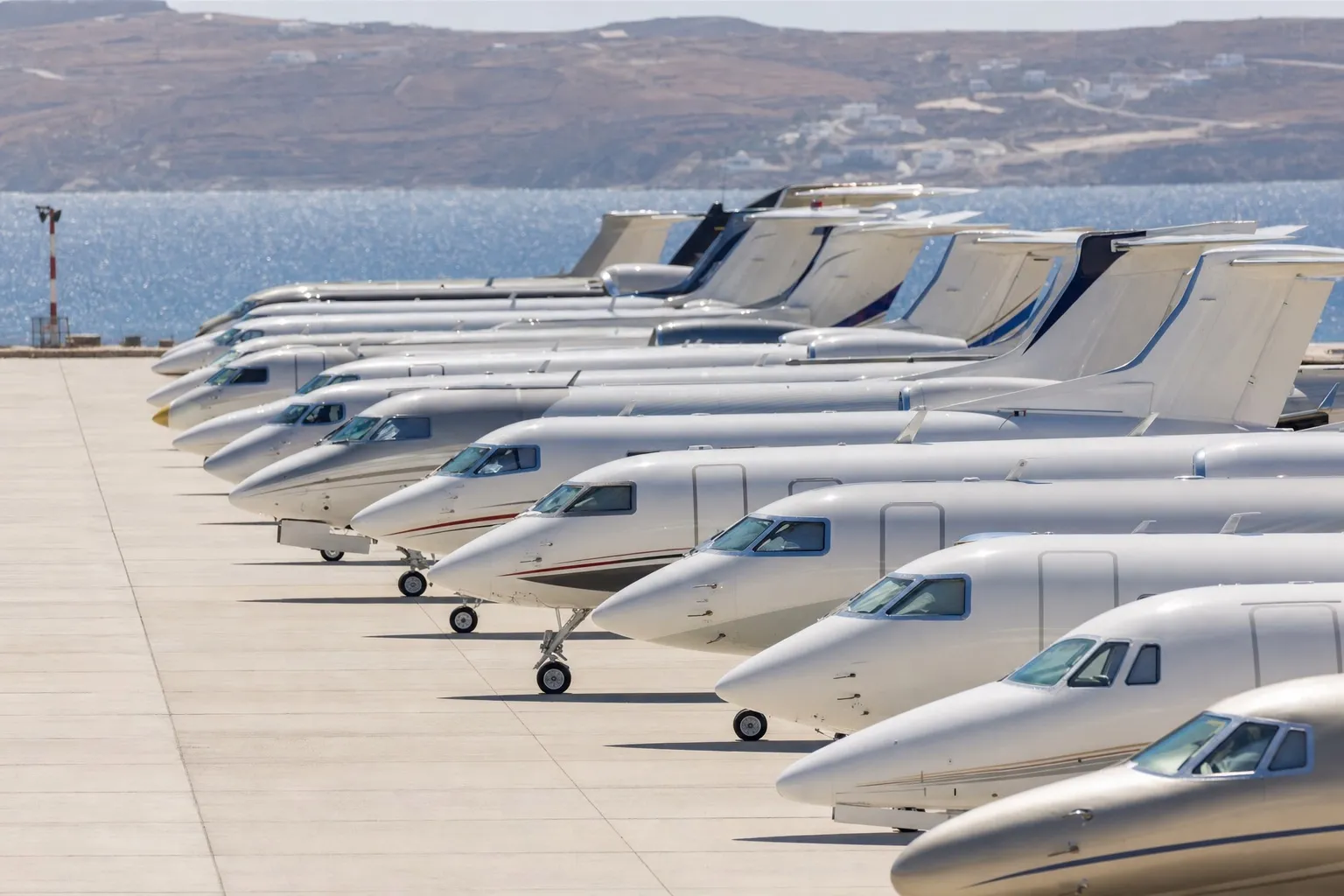 Private jets parked wingtip to wingtip on a sun-drenched Mediterranean airport apron with blue sea and hills in the background