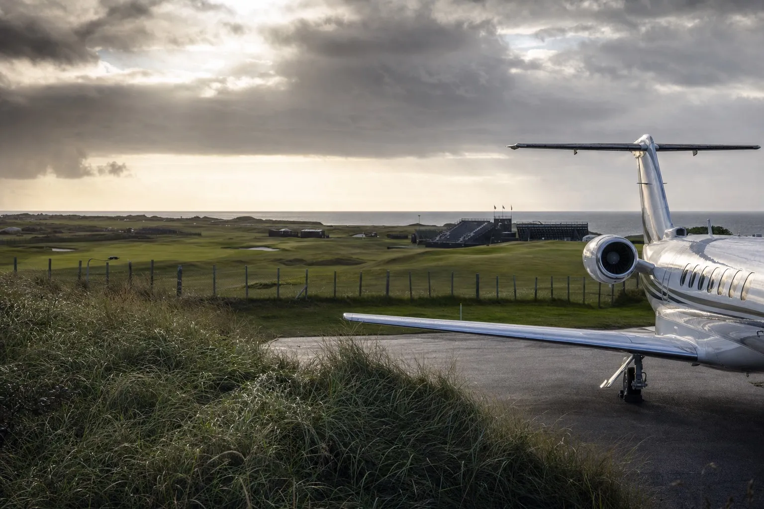 A light jet parked at a small coastal airfield with links golf dunes and a grey-blue sea in the background
